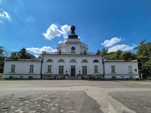 Pałac w Jabłonnie, biała, klasycystyczna budowla z charakterystyczną kopułą, otoczona zielenią, pod błękitnym niebem z chmurami.