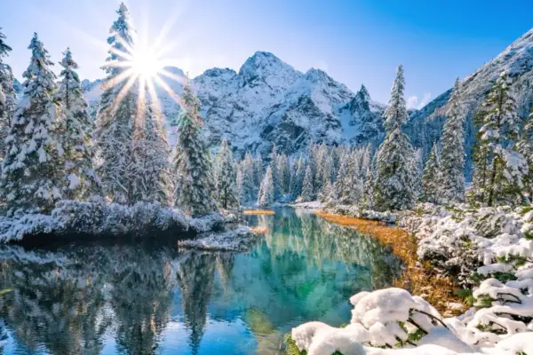 Morskie Oko zimą, otoczone ośnieżonymi świerkami i górami, odbija się w turkusowej tafli wody. Słońce przebija się przez drzewa, tworząc promienie.