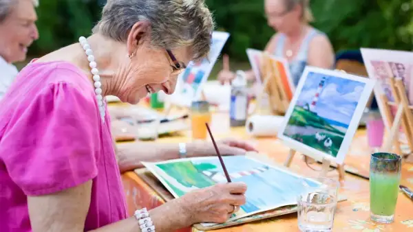 Des personnes âgées participent à des activités manuelles en peignant en plein air. Une dame souriante, vêtue d'un haut rose, est concentrée sur sa toile.