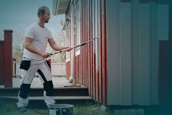Man målar om huset med en lång roller. Den röda träfasaden får nya grå paneler.