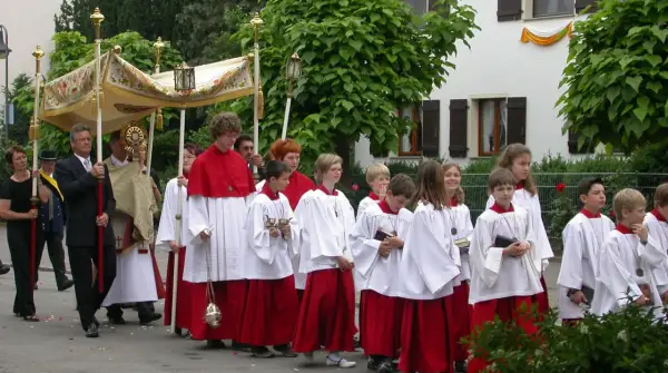 Prozession am Corpus Christi Feiertag mit Ministranten in weißen und roten Gewändern, die eine Monstranz tragen.