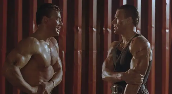 Dwaj Jean-Claude Van Damme w scenie z jednego z jego filmów, spoceni i z widocznymi ranami, patrzą na siebie.