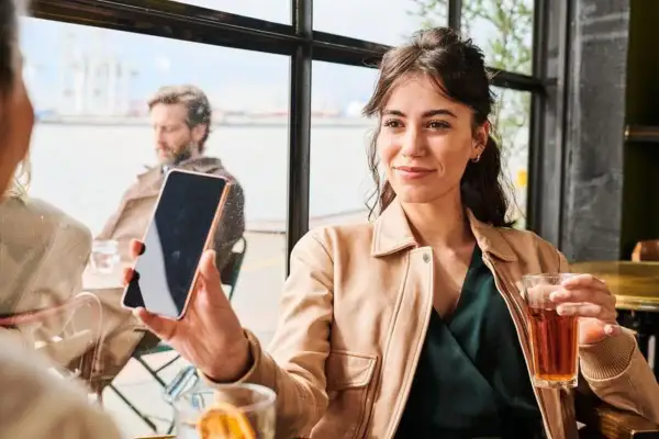 Kobieta trzyma telefon, słuchając wiadomości na poczcie głosowej. W tle mężczyzna i widok na port.