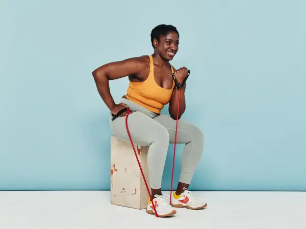 Woman smiles while performing arm strength exercises with a resistance band, seated on a box.