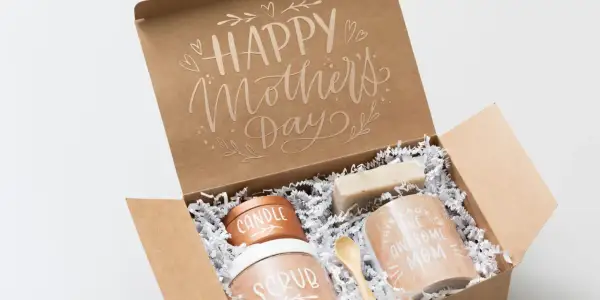 Otwarty brązowy karton z napisem "Happy Mother's Day" i zawartością: świeca, peeling, mydło, kubek i drewniana łyżeczka.