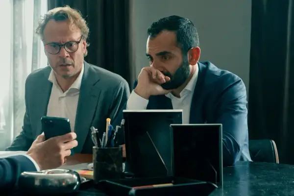Två män i kostym sitter vid ett bord. Den ena mannen med glasögon tittar på sin mobiltelefon, den andra mannen funderar. Scen från "Hundarna" på SVT.