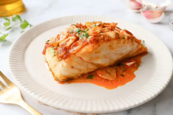 Delicioso bacalao a la llauna con salsa de tomate y ajo, servido en un plato blanco con un tenedor dorado.