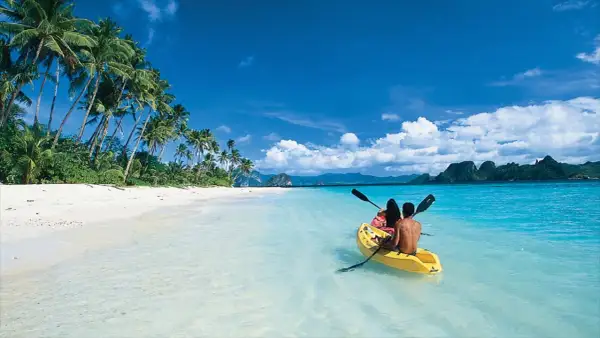 Två personer paddlar kajak i det turkosa vattnet vid en vit sandstrand med palmer på Palawan, Filippinerna.