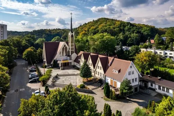 Nowoczesny kościół z wieżą i przylegającym budynkiem parafialnym, otoczony zielenią. W oddali widać wzgórza i zabudowania. To miejsce, gdzie historia spotyka się z teraźniejszością, jak w vii dwór.