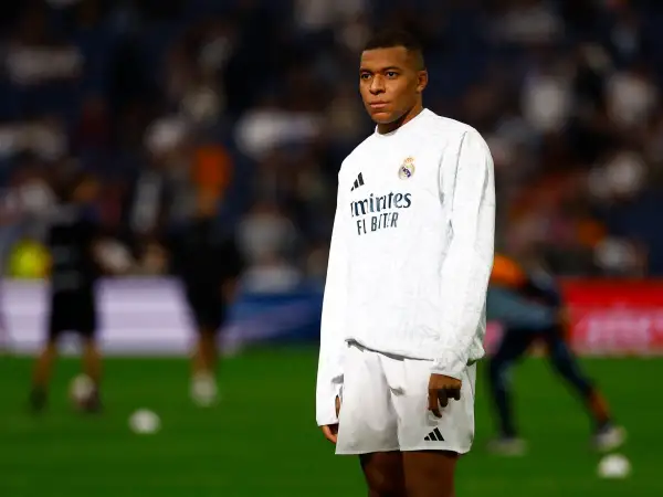 Mbappé, młody mbp piłkarz, w białej koszulce i spodenkach Realu Madryt, stoi na murawie stadionu.