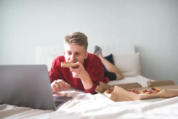 A young man eats pizza in bed while watching a laptop. Is it bad to go to sleep after eating? This scene suggests a late-night snack before bed.