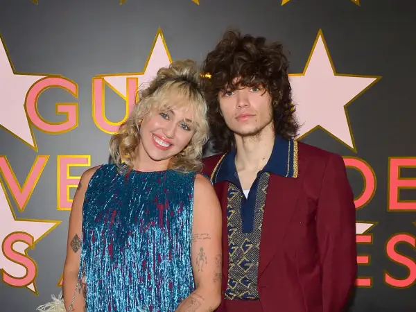 Miley Cyrus y Max Morando posan sonrientes. Ella lleva un vestido azul brillante con flecos, él un traje burdeos con detalles dorados.