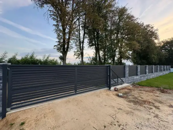 Nowe, nowoczesne ogrodzenie z ciemnoszarymi panelami i kamiennymi słupkami. W tle widać wysokie drzewa w linii ogrodzenia, tworzące naturalną barierę.