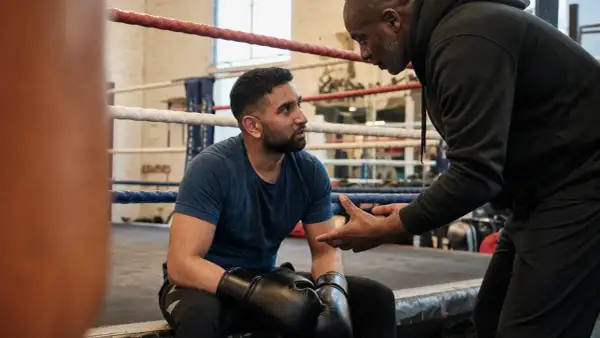 A boxer, sweat on his brow, listens intently as his coach explains that boxing is hard, but worth it.