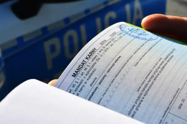 Dłoń trzyma mandat karny, dokument potwierdzający popełnienie wykroczenia. W tle widoczny napis "POLICJA".