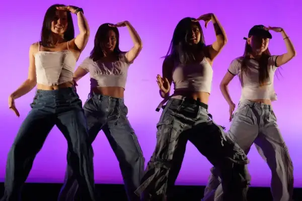 Cztery tancerki w luźnych jeansach i krótkich topach wykonują choreografię w stylu **key pop**.