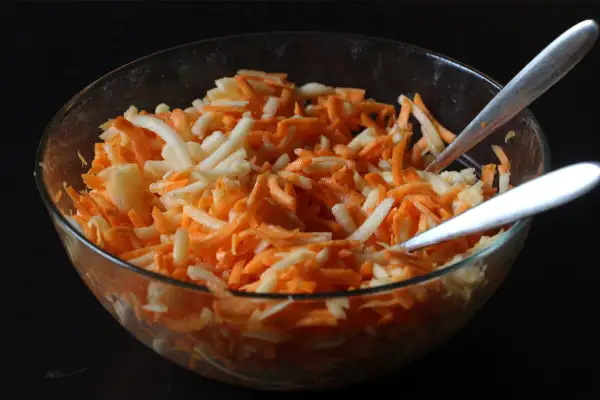 Schüssel mit geriebenen Karotten und Äpfeln, bereit für ein leckeres karottensalat rezept. Zwei Löffel warten darauf, den Salat zu servieren.