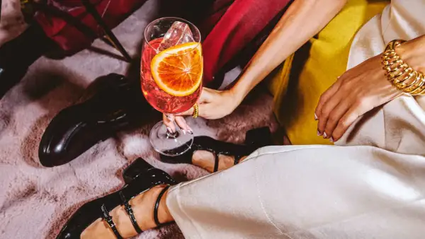 Elegancka dłoń trzyma kieliszek z drinkiem, w którym króluje wytrawny wermut. Obok błyszczą czarne buty i złota biżuteria.