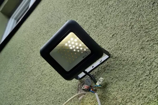 Czarny reflektor LED zamontowany na zielonej ścianie. Żarzące się ledy pobierają prąd, co widać po podłączonych przewodach.