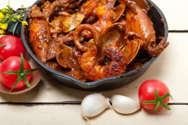 Un plat de calamars à l'armoricaine mijoté dans une sauce rouge, servi dans une poêle en fonte, avec des tomates et de l'ail à proximité.