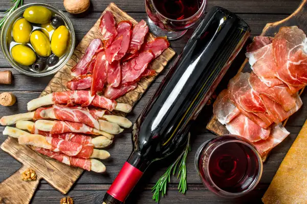 Un bicchiere di vino rosso, una bottiglia, salumi, olive e grissini avvolti nel prosciutto, un'ambientazione perfetta per la vinificazione in rosso.