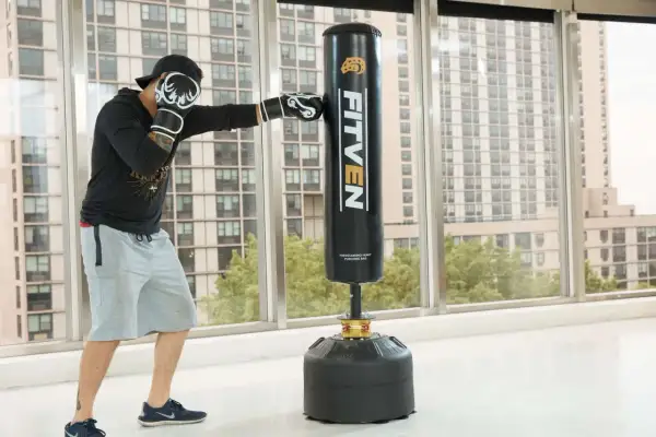 Man in boxing gloves practices how to punch a punching bag in a bright room with a city view.