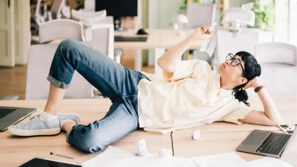 En person ligger utsträckt på ett skrivbord, omgiven av papper och laptops. Klädd i jeans och en ljus skjorta, ser hen ut att fundera över en papperslapp, en bild av en dysfunktionell arbetsplats.