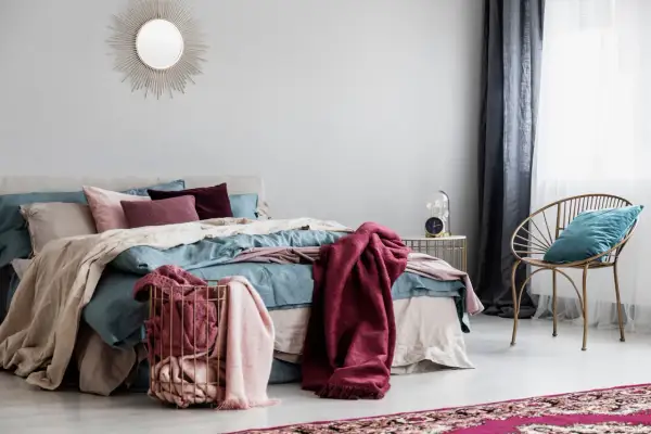 Letto con coperte color borgogna e cuscini, un cesto dorato con plaid rosa e una sedia dorata con cuscino turchese.