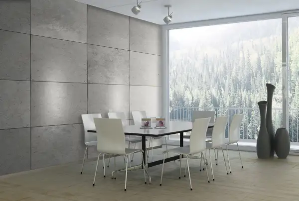 Esszimmer mit moderner Beton Optik Wandverkleidung, weißem Tisch und Stühlen, sowie einem Panoramafenster mit Blick auf einen Wald.