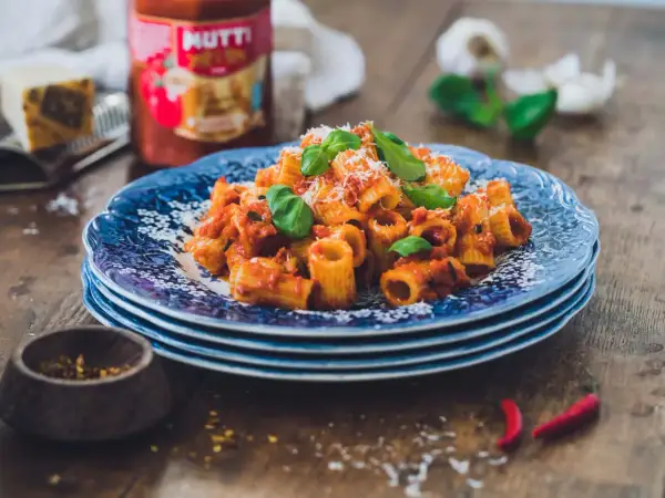 En tallrik med rigatoni all'arrabiata, toppad med basilika och parmesan. Bredvid syns en burk Mutti-tomatsås, vitlök och chilifrukter.