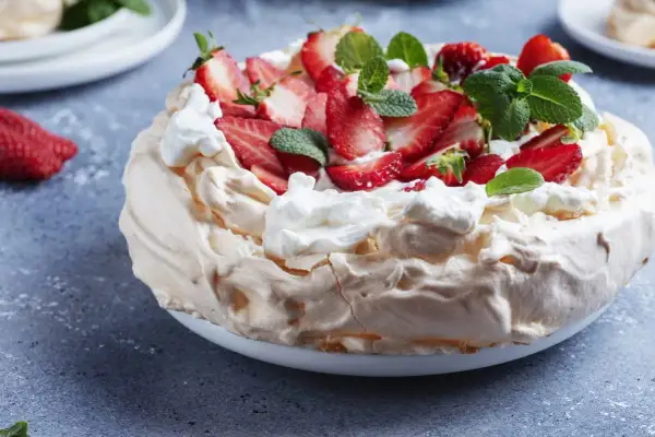 Pyszny tort bezowy z mascarpone i bitą śmietaną, ozdobiony świeżymi truskawkami i miętą.