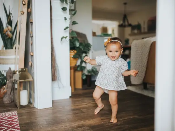 Radosne dziecko w kwiecistej sukience robi pierwsze kroki, kiedy dziecko zaczyna wstawać. W tle domowe wnętrze z roślinami.