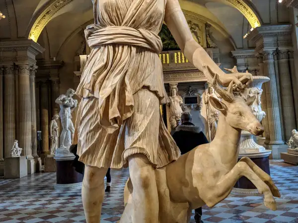 Statue de Diane chasseresse, qui est Artémis, tenant un cerf. L'œuvre est dans un musée, avec d'autres sculptures en arrière-plan.