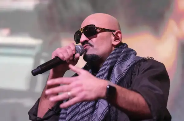 Ein bekannter Rapper mit Sonnenbrille und Schal spricht ins Mikrofon, während er gestikuliert.