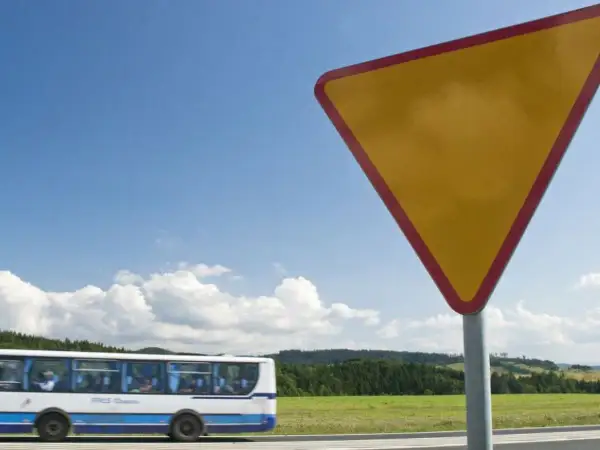 Żółty trójkątny znak a7 ostrzega przed ustąpieniem pierwszeństwa. W tle jedzie autobus, a za nim zielone wzgórza.