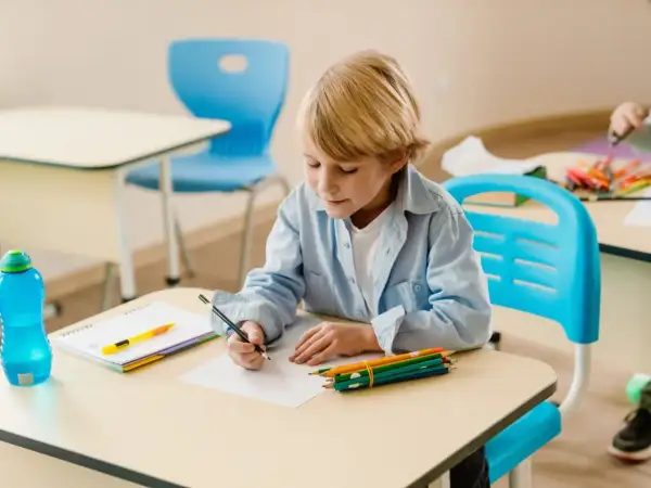 Chłopiec z blond włosami, ubrany w błękitną koszulę, siedzi przy biurku i rysuje. Obok niego leżą kredki, zeszyt i butelka z wodą. Coraz lepiej mu idzie.