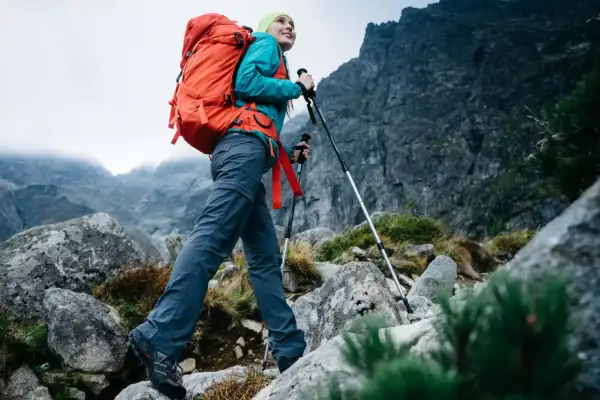 Kobieta z plecakiem wyrusza na wycieczkę w góry. Używa kijków trekkingowych, by pokonać kamienisty szlak.