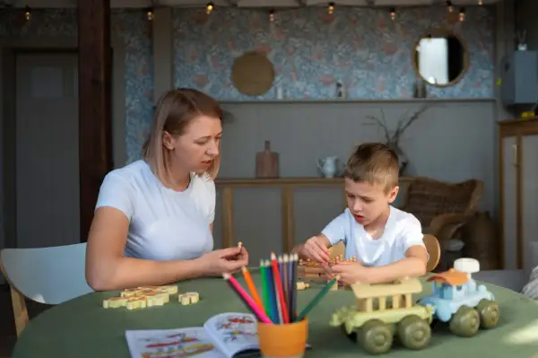 Kobieta i chłopiec grają w drewniane gry na zielonym stole. W tle widać wystrój pokoju, przypominający salę w przedszkolu niepublicznym.