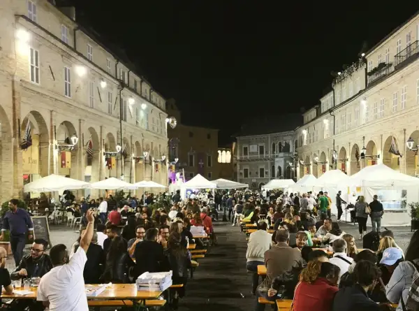 Serata di festa in una piazza delle Marche, con tavoli all'aperto e gente che mangia e socializza.