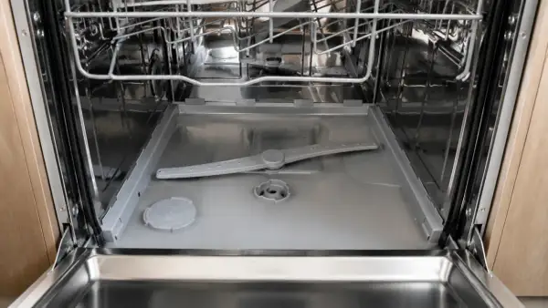 Intérieur d'un lave-vaisselle ouvert, montrant le bras d'aspersion et le fond. On observe de l'eau stagnante au niveau du filtre, signe d'un nettoyage nécessaire.