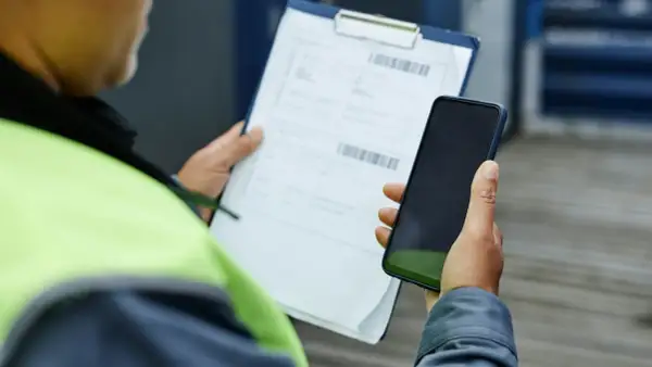 Pracownik w kamizelce odblaskowej trzyma telefon i dokumenty. Zastanawia się, czy warto złożyć skargę do PIP.