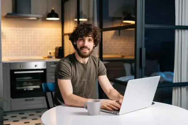 Uśmiechnięty mężczyzna z kręconymi włosami pracuje na laptopie w kuchni. Myśli o tym, jak zapłacić czynsz, ale jest zrelaksowany.