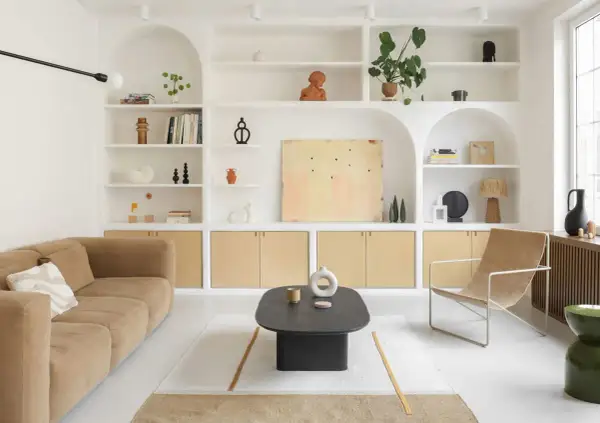 Salon épuré avec canapé beige, fauteuil en toile, table basse noire et étagères murales remplies d'objets décoratifs. Le tout baigne dans une douce lumière, rehaussée par la **couleur blanc cassé** des murs et des étagères.