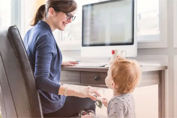 En kvinna med glasögon ler mot sitt barn vid ett skrivbord. Hon visar vad betyder flexibel genom att balansera arbete och familjeliv.