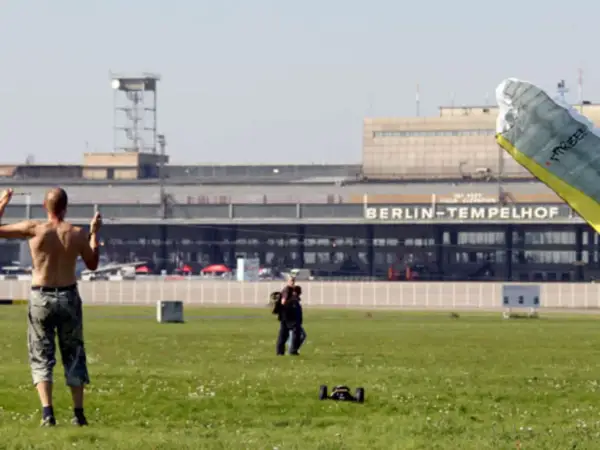 Ein Mann mit nacktem Oberkörper steht auf dem Feld vor dem BERLIN-TEMPELHOF. Ein Gleitschirm liegt im Vordergrund.