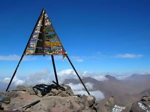 Sommet du mont Toubkal, altitude impressionnante. Le cairn triangulaire, couvert de graffitis, se dresse fièrement sur un pic rocheux, dominant un paysage montagneux spectaculaire sous un ciel bleu éclatant.