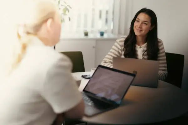 Två kvinnor sitter vid ett bord med laptops. En kvinna med mörkt hår ler och tittar bort, kanske under en intervju med chefen.