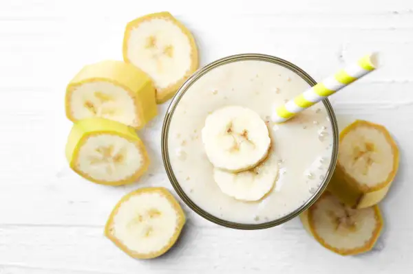 Un cremoso frullato di frutta con latte, guarnito con fette di banana e una cannuccia a righe gialle e bianche, circondato da fette di banana fresca.