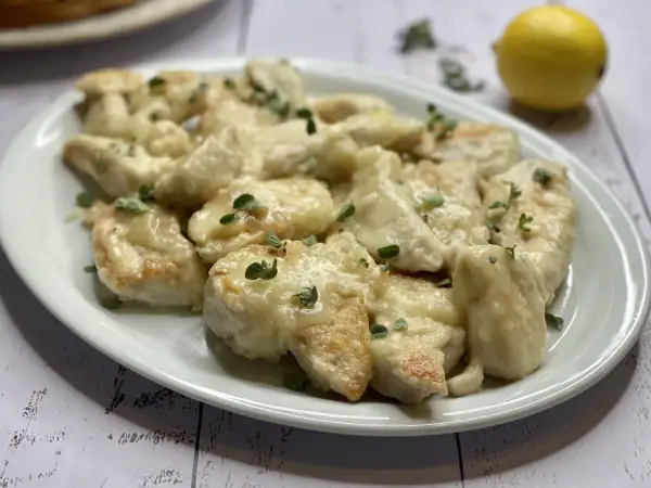 Delizioso pollo al limone ricetta, servito su un piatto bianco con erbe aromatiche e un limone sullo sfondo.