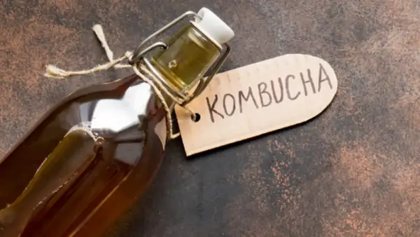En flaska med en mörk, bubblig kombucha dryck. En brun etikett med texten "Kombucha" hänger på flaskhalsen.